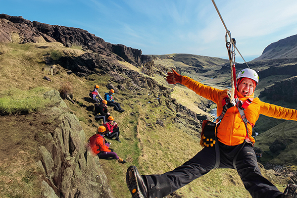 A woman ziplines in Vík, Iceland, on a spring day with a blue sky and spectators watching on a grassy slope.